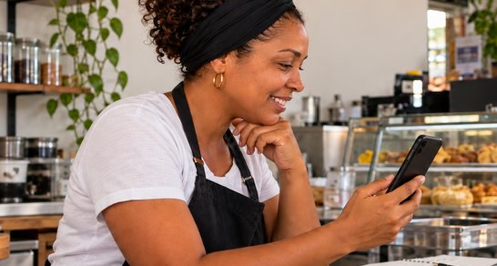 Empreendedora sorrindo enquanto usa o celular em seu comércio